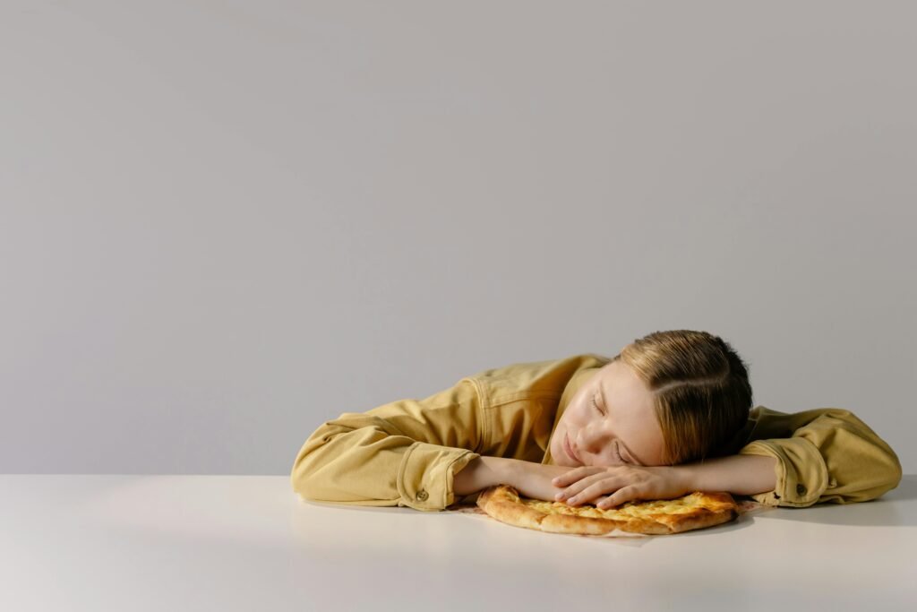 A Caucasian woman with eyes closed, resting her head on a pizza, embodying relaxation and humor.